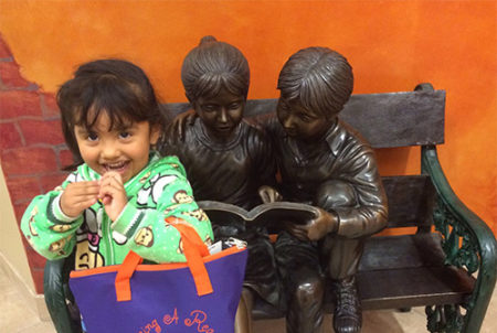 child holding purple bag next to statue of children on a bench