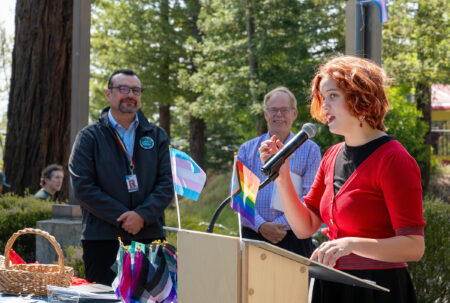 A LGBTQ+ Student stands outside at a podium talking to a crowd of onlookers about the Rainbow flag raising